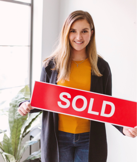 A woman holding a sold sign in front of a window.
